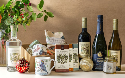 A curated gift assortment featuring Waddeison-branded items including a gin bottle, dark chocolate bars, a wine bottle, a candle jar, a ceramic bird mug, and a small decorative box, arranged on a tabletop with floral and holiday ornaments against a warm neutral backdrop.